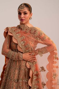 Pakistani Bridal Dress in Peach Lehenga Pishwas Style is an embellished attire that will give you a head-turning look on the most important day of your life. Lavish designs, embroidery, and ornaments give a chic look to this beautiful Pishwas Dress. Pakistani Bridal Pishwas: Pakistani Bridal Pishwas in an alluring peach shade has premium net fabric. The Pishwas Frock is gracefully emblazoned with zardosi, dabka, floral designs, and naqshi. The Traditional Pishwas is embellished with intricate designs, silver and gold details, giving it a flawless finishing look. Bridal Lehenga: The Bridal Lehenga has premium quality tissue fabric and it looks perfect with the heavily embellished Pishwas Frock. The Lehenga in peach gold color has a huge flare and its borders are emblazoned with tilla, naqsh Pishwas Pakistani, Bridal Pishwas, Bridal Lehenga Pakistani, Pakistani Bridal Lehenga, Peach Lehenga, Tissue Fabric, Gold Lehenga, Pakistani Bridal Dress, Dress Pakistani