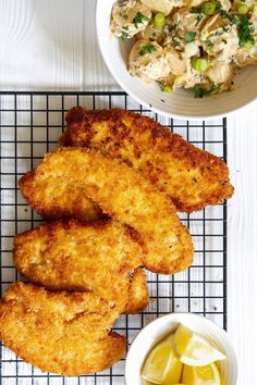 some fried chicken on a wire rack next to a bowl of sauce and lemon wedges