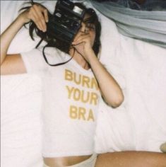 a woman laying in bed with a camera and t - shirt that says burn your bra