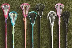 four different colored lacrosse sticks lined up in a row on the ground with grass behind them