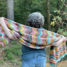 a woman is holding up a multicolored knitted shawl in the woods