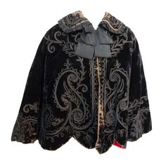 Edwardian black velvet cape with soutache trim on collar and throughout body. Silk neck bow. Hook and eye closure along front. Re-lined in red cotton Luxury Black Medieval Outerwear, Luxury Medieval Black Outerwear, Luxury Medieval Style Black Outerwear, Luxury Black Gothic Cape, Elegant Velvet Outerwear For Costume, Black Velvet Costume Outerwear, Velvet Cape, Black Cape, Capes For Women