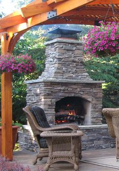 an outdoor fireplace with wicker chairs under it and flowers hanging from the pergolated roof