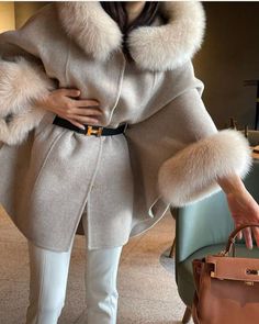 a woman wearing a fur coat and white pants holding a brown handbag while standing next to a purse