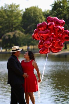 a man and woman standing next to each other holding red heart balloons in front of water