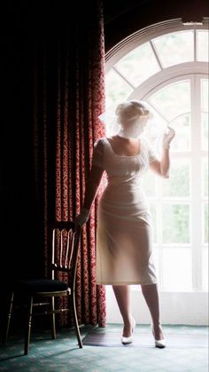 a woman standing in front of a window wearing a white dress and holding a hat