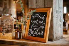 This shows a bar at a wedding and the display on it. On the bar shows different sized and coloured stoneware bottle vases in it are some greenery. Next to the bottles is a framed sign that reads "waste not want not, please reuse your glass" The sign is resting on a vintage crate. Display Board