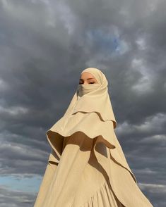 a woman in a white dress and head scarf is standing on the beach under a cloudy sky