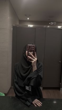 a woman taking a selfie with her cell phone in a public restroom, wearing a black hijab