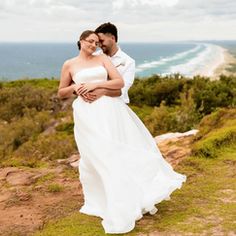 a man and woman standing on top of a hill next to the ocean with their arms around each other