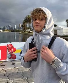 a young man standing next to a body of water holding a cell phone and wearing a hoodie