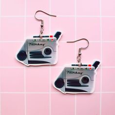 two pairs of earrings with an old school radio cassette player on them, hanging from hooks in front of a pink tiled wall