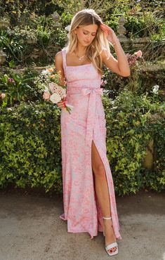 a woman standing in front of bushes wearing a pink floral print dress with thigh high slit