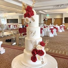 a white wedding cake with red flowers on top in a banquet room decorated with tables and chairs