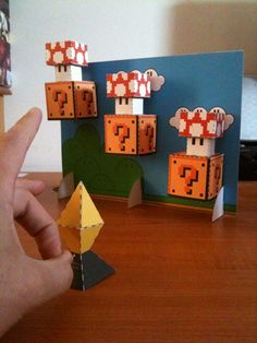 a hand is holding an origami piece in front of some paper mario mushroom structures