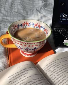 an open book is next to a bowl of soup and a laptop on a bed