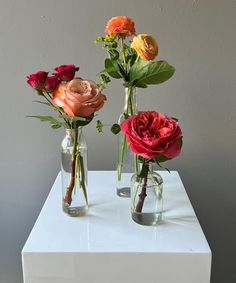 three vases filled with different colored flowers on top of a white table next to a gray wall