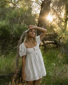 a woman in a white dress is standing by a tree with her hands on her head