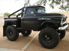 an old black truck with big tires parked in a parking lot next to a tree