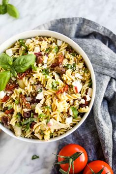 a white bowl filled with pasta salad next to two tomatoes and basil leaves on the side