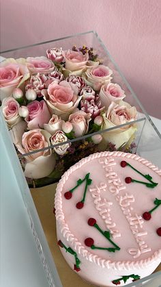 a birthday cake and bouquet of roses in a clear box on a table next to a pink wall