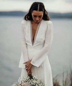 a woman in a white dress holding a bouquet of flowers near the water's edge