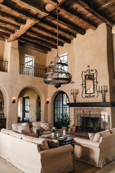 Rustic living room with high ceilings, wooden beams, a chandelier, beige sofas, and an ornate fireplace. Spanish Art Deco Interior, Spanish Style Hacienda Home, Lime Paint Interior, Modern Spanish Style Homes Living Room, Traditional Colonial House Interior, Modern Spanish House Interior Design, Spanish Farmhouse Interior, Old Spanish House Interior, Modern Heritage Home