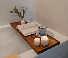 two candles are sitting on a wooden tray in front of a bathtub with an open book