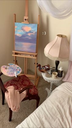 an easel with a painting on it next to a chair and lamp in a bedroom