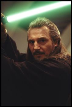 a man with long hair holding a green light saber in his right hand and looking at the camera