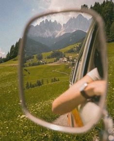 a side view mirror with mountains in the back ground and green grass on both sides