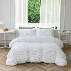 a bed with white comforter and pillows in a room next to a window that has wood flooring