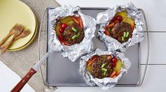 four foil wrapped hamburgers sitting on top of a pan with meat and veggies