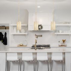 a white kitchen with gold accents and brass pendant lights hanging over the island, along with four bar stools