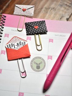 an open notebook with paper clips and magnets attached to it next to a pink pen