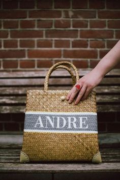 a person's hand holding onto a woven bag on a wooden bench in front of a brick wall