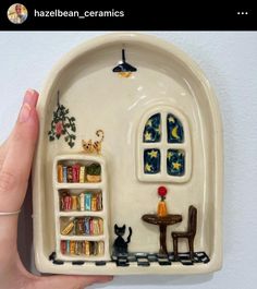 a hand holding up a ceramic plate with a miniature house on the front and bookshelf in the back