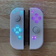 two video game controllers sitting next to each other on a wooden surface with the lights turned on