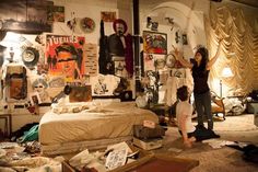 a woman and child are standing in a messy room with posters on the wall behind them