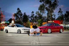 two cars parked next to each other in a parking lot with people sitting on the ground