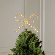 a christmas tree decorated with lights and a gold bow on it's top, in front of a white wall