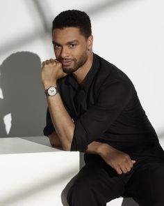 a man in black shirt and pants sitting on a white wall with his hand on his chin