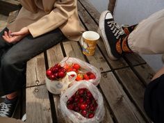 two people sitting on a wooden bench next to baskets of cherries and coffee cups