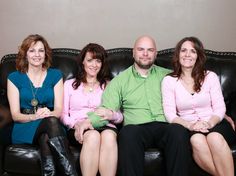 four people sitting on a black couch smiling for the camera, with one woman in pink and two men in green