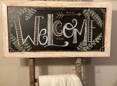 a welcome sign hanging from the side of a wooden frame on a towel rack in a bathroom