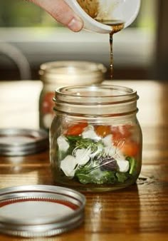 a person pouring dressing into a mason jar filled with vegetables and spinach, on top of a wooden table