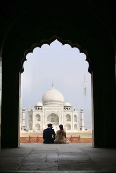 two people sitting on the ground in front of a building with an archway leading to it