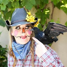 a woman with painted face and bird on her head pointing to the side while wearing a scarecrow hat