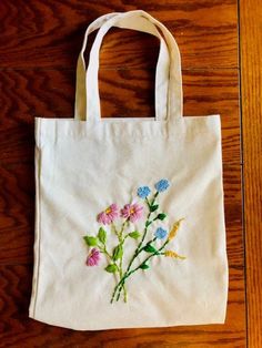 a white tote bag with flowers embroidered on the front and side, sitting on a wooden floor