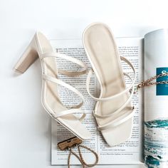 A pair of ivory strappy heels that have a square toe. These shoes are pictured on a white background with gold jewelry and a magazine. Cream Ankle Strap Block Heels For Summer, Cream High Heel Block Heels For Summer, Cream Block Heels With Stacked Heel For Summer, Summer Cream Block Heels With Stacked Heel, Stacked High Heel Shoes For Day Out, Block Heels With Wrapped Heel For Day Out, Stacked Heel High Heels For Day Out, Wrapped Block Heel For Day Out, Cream Heels For Summer Day Out